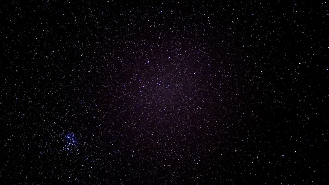 A starry night sky filled with countless tiny specks of light spread across a dark canvas. In the bottom left, a cluster of brighter stars stands out, forming a distinct grouping.