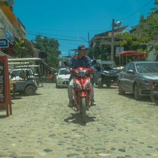 A person is riding a red scooter down a cobblestone street lined with parked vehicles, including cars and golf carts. The street is in a small town, with various storefronts and signs visible. Trees and utility poles are in the background under a clear blue sky.