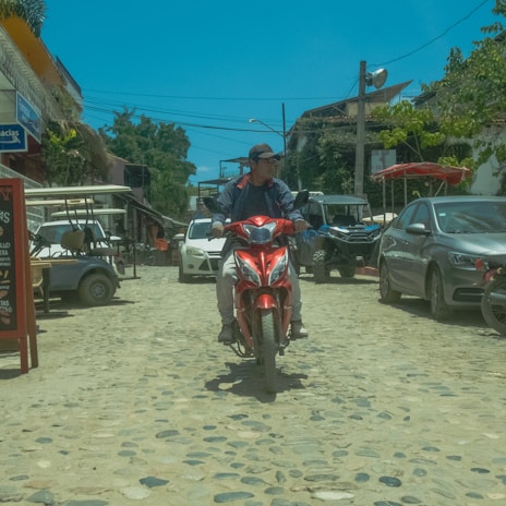 A person is riding a red scooter down a cobblestone street lined with parked vehicles, including cars and golf carts. The street is in a small town, with various storefronts and signs visible. Trees and utility poles are in the background under a clear blue sky.