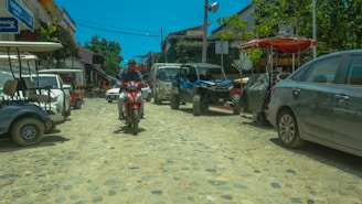 A cobblestone street lined with various parked vehicles, including golf carts, a motorcycle, and an all-terrain vehicle. The street is bordered by shops and trees, with clear blue skies overhead. There is a person riding a red motorcycle down the middle of the street.