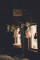 A small shop with a sign reading 'Old Town Gifts' is illuminated by soft lighting. Several Swedish flags hang above the entrance, alongside a display of tote bags with various designs. Baskets filled with goods are placed near the entrance, visible through the shop's glass window that reveals a warmly lit interior.