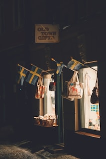 A small shop with a sign reading 'Old Town Gifts' is illuminated by soft lighting. Several Swedish flags hang above the entrance, alongside a display of tote bags with various designs. Baskets filled with goods are placed near the entrance, visible through the shop's glass window that reveals a warmly lit interior.