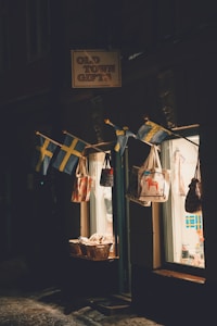 A small shop with a sign reading 'Old Town Gifts' is illuminated by soft lighting. Several Swedish flags hang above the entrance, alongside a display of tote bags with various designs. Baskets filled with goods are placed near the entrance, visible through the shop's glass window that reveals a warmly lit interior.