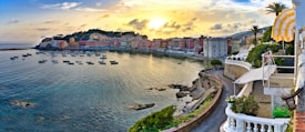 A picturesque seaside town features colorful buildings lining a curved shoreline under a golden sunset. Small boats are moored in the calm water, and a winding path with lush greenery leads along the coast. The vibrant sky is reflected in the sea, creating a serene and inviting atmosphere.