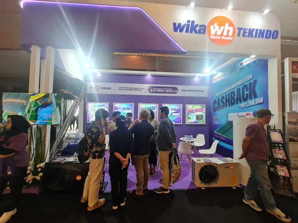 A booth at a trade exhibition with several people gathered around looking at promotional information. The booth features displays and a seating area, surrounded by vibrant advertisements with text about discounts and offers related to water heaters.