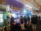 A trade show booth features a large video display screen showing a worker in a yellow helmet and safety gear. Several people are gathered around the booth, engaging with promotional materials and displays that include various posters and banners. The booth is well-lit with overhead lighting, and there are plants decorating the space.