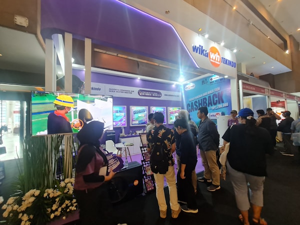 A trade show booth features a large video display screen showing a worker in a yellow helmet and safety gear. Several people are gathered around the booth, engaging with promotional materials and displays that include various posters and banners. The booth is well-lit with overhead lighting, and there are plants decorating the space.
