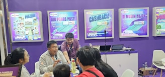 A group of people is engaged in conversation at an exhibition booth. The booth has purple walls with several promotional posters displayed, advertising various products. A table is set up with laptops and promotional materials, and the people appear to be attentively discussing or presenting something related to the exhibits.