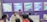 A group of people is engaged in conversation at an exhibition booth. The booth has purple walls with several promotional posters displayed, advertising various products. A table is set up with laptops and promotional materials, and the people appear to be attentively discussing or presenting something related to the exhibits.