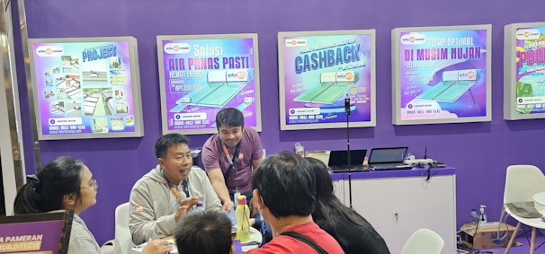 A group of people is engaged in conversation at an exhibition booth. The booth has purple walls with several promotional posters displayed, advertising various products. A table is set up with laptops and promotional materials, and the people appear to be attentively discussing or presenting something related to the exhibits.