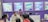 A group of people is engaged in conversation at an exhibition booth. The booth has purple walls with several promotional posters displayed, advertising various products. A table is set up with laptops and promotional materials, and the people appear to be attentively discussing or presenting something related to the exhibits.