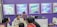 A group of people is engaged in conversation at an exhibition booth. The booth has purple walls with several promotional posters displayed, advertising various products. A table is set up with laptops and promotional materials, and the people appear to be attentively discussing or presenting something related to the exhibits.