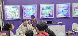 A group of people is engaged in conversation at an exhibition booth. The booth has purple walls with several promotional posters displayed, advertising various products. A table is set up with laptops and promotional materials, and the people appear to be attentively discussing or presenting something related to the exhibits.