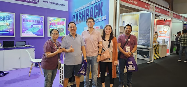 A group of five people stands together smiling at a trade show or exhibition. They are dressed casually, and three of them hold bags with promotional text. Behind them, there are colorful banners and booths with promotional signage and products on display. One booth has electronic equipment and screens showing various promotions.