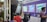 A group of people is gathered around a promotional booth within an indoor exhibition or trade show setting. There are posters advertising promotional offers and business branding on the purple wall behind them. A man is standing to the right, possibly giving a presentation or information session.