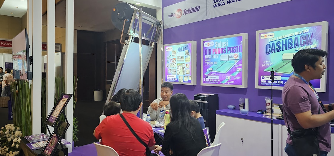 A group of people is gathered around a promotional booth within an indoor exhibition or trade show setting. There are posters advertising promotional offers and business branding on the purple wall behind them. A man is standing to the right, possibly giving a presentation or information session.