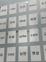 A collection of white cards attached to a light gray wall, each featuring a single word in either English or Korean. The words are printed in black, with some examples including 'vibe', 'slow', 'wave', and 'dream' in English and corresponding Korean words.