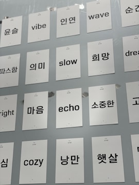 A collection of white cards attached to a light gray wall, each featuring a single word in either English or Korean. The words are printed in black, with some examples including 'vibe', 'slow', 'wave', and 'dream' in English and corresponding Korean words.