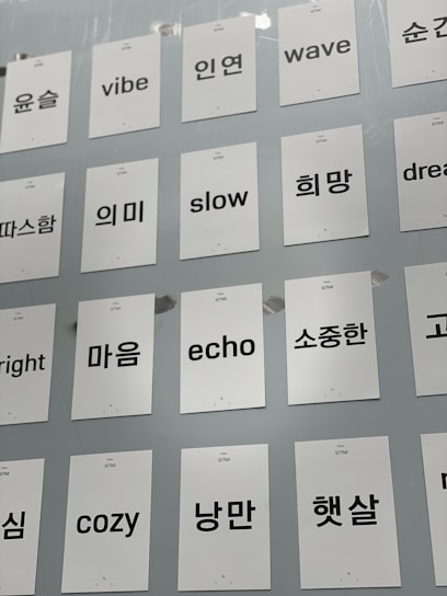 A collection of white cards attached to a light gray wall, each featuring a single word in either English or Korean. The words are printed in black, with some examples including 'vibe', 'slow', 'wave', and 'dream' in English and corresponding Korean words.