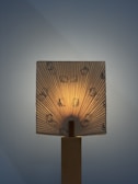 A lamp with a fabric or paper shade featuring cartoon characters is illuminated from behind. The light creates a warm glow and highlights the vertical pleats of the shade. The base appears to be made of wood, adding a natural element to the design.