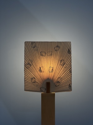 A lamp with a fabric or paper shade featuring cartoon characters is illuminated from behind. The light creates a warm glow and highlights the vertical pleats of the shade. The base appears to be made of wood, adding a natural element to the design.