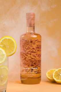 A beautifully designed bottle of rare citrus gin with intricate floral patterns sits against a warm, smoky background. A glass of water garnished with lemon slices and two halved lemons are placed next to the bottle, adding a refreshing citrus element.