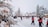 A winter scene with snow-covered trees and a festive atmosphere. People are walking around in winter clothing, surrounded by decorative lights and snowy landscape. Buildings with pointed roofs are visible in the background, enhancing the wintry feel.