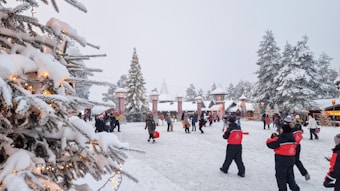 A winter scene with snow-covered trees and a festive atmosphere. People are walking around in winter clothing, surrounded by decorative lights and snowy landscape. Buildings with pointed roofs are visible in the background, enhancing the wintry feel.