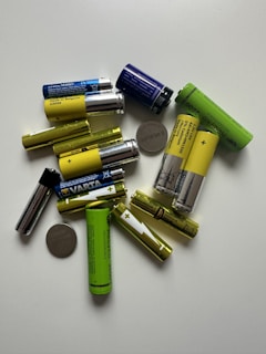 A collection of various batteries of different sizes and colors scattered on a white surface. The batteries include cylindrical AA or AAA types in yellow, green, and blue, as well as flat, coin-shaped ones.
