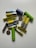 A collection of various batteries of different sizes and colors scattered on a white surface. The batteries include cylindrical AA or AAA types in yellow, green, and blue, as well as flat, coin-shaped ones.