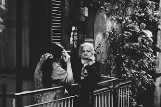 Two elderly individuals are standing on a balcony that overlooks a street. One person is facing towards the balcony railing, while the other is engaged in a conversation and appears to be gesturing. The background includes a partially visible window with shutters, a wall with some wear and tear, and foliage.