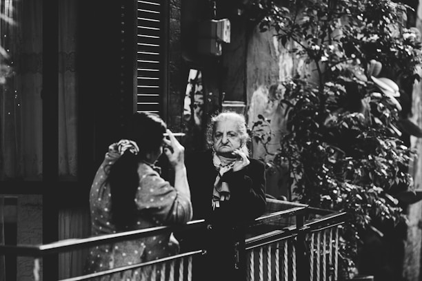 Two elderly individuals are standing on a balcony that overlooks a street. One person is facing towards the balcony railing, while the other is engaged in a conversation and appears to be gesturing. The background includes a partially visible window with shutters, a wall with some wear and tear, and foliage.