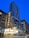A tall modern skyscraper towers above a classic theater marquee with bright, illuminated signage bearing the name 'Chicago'. The streetscape features a blend of historical and contemporary architecture with pedestrians and a parked bicycle. The sky is a vivid blue, suggesting early evening.