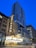 A tall modern skyscraper towers above a classic theater marquee with bright, illuminated signage bearing the name 'Chicago'. The streetscape features a blend of historical and contemporary architecture with pedestrians and a parked bicycle. The sky is a vivid blue, suggesting early evening.