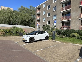 A white car is parked in a residential parking spot. The car is plugged into an electric vehicle charging station. The parking area is paved with brick and is situated next to a building with several floors and balconies. There are some plants and shrubs surrounding the area, and tall trees can be seen in the background under a clear blue sky.