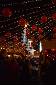 A festive outdoor scene with strings of lights and large red ornaments hanging overhead. There is a crowd of people gathered below, engaged in various activities. In the background, there is a brightly lit tree and some market stalls.