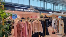 A stylish store interior with racks of clothing displayed under decorative structures and plants. The racks feature a variety of garments such as jackets and dresses in soft pastel and neutral tones. Signage for a 'second hand & caf&eacute;' is visible, suggesting a combination of retail and dining space. The ambiance is modern and welcoming, enhanced by natural lighting from large windows.