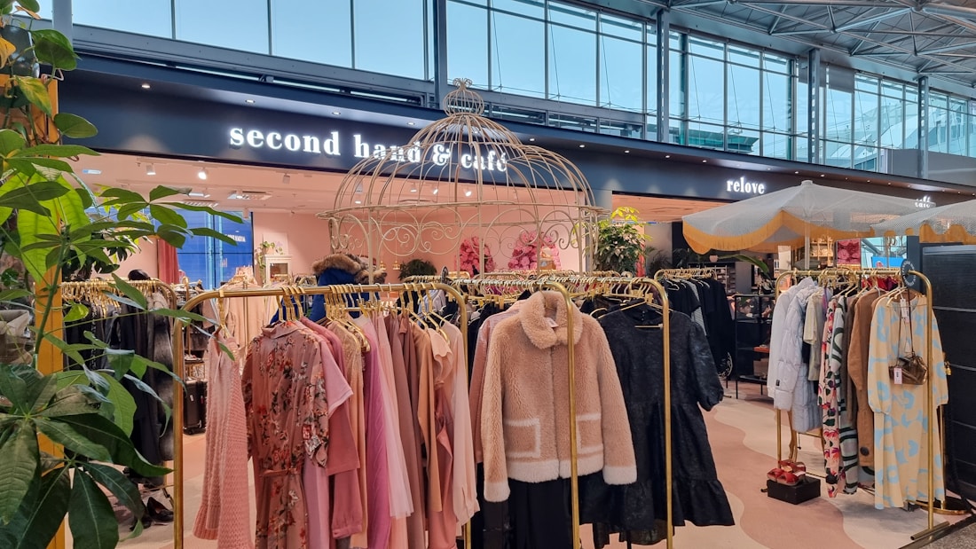 A stylish store interior with racks of clothing displayed under decorative structures and plants. The racks feature a variety of garments such as jackets and dresses in soft pastel and neutral tones. Signage for a 'second hand & caf&eacute;' is visible, suggesting a combination of retail and dining space. The ambiance is modern and welcoming, enhanced by natural lighting from large windows.