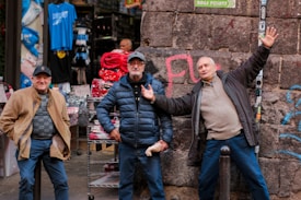 Three men are standing and posing in front of an outdoor shop display with clothing and various items. They are casually dressed, and one of them is gesturing energetically. Behind them is a stone wall with graffiti, and the shop has racks displaying shirts in different colors.