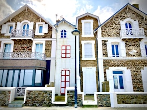 Three attached houses with a rustic architectural style featuring stone exteriors and white window shutters. The middle house has red doors and windows, while the houses on the sides have more uniform designs with balconies. A street lamp stands in front of the middle house.