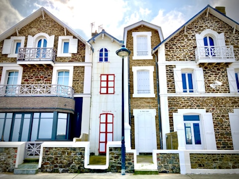 Three attached houses with a rustic architectural style featuring stone exteriors and white window shutters. The middle house has red doors and windows, while the houses on the sides have more uniform designs with balconies. A street lamp stands in front of the middle house.