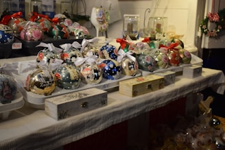A collection of intricately decorated Christmas ornaments displayed on a table, featuring various designs such as flowers, cityscapes, and patterns. Some ornaments have red or white ribbons attached. The table is covered with a light-colored cloth, and several small decorative boxes are placed alongside the ornaments. The background includes additional decorations and festive elements.