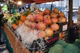 A market stall displays an array of fresh produce with pomegranates stacked in boxes lined with straw. Various fruits including oranges and what appear to be pears are in the background, set under a canopy. A person is partially visible to the side, seemingly interacting with the stall.