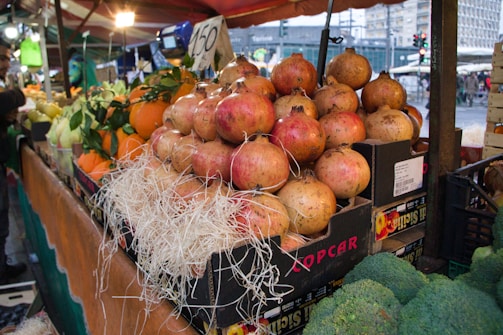 A market stall displays an array of fresh produce with pomegranates stacked in boxes lined with straw. Various fruits including oranges and what appear to be pears are in the background, set under a canopy. A person is partially visible to the side, seemingly interacting with the stall.