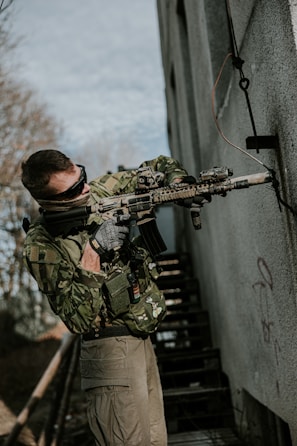 A person dressed in camouflage military gear holds a rifle while standing next to a building with graffiti. They are wearing tactical gloves and sunglasses, with a patch on their vest. The scene appears to be outdoors near a staircase and some foliage.