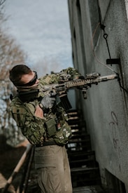 A person dressed in camouflage military gear holds a rifle while standing next to a building with graffiti. They are wearing tactical gloves and sunglasses, with a patch on their vest. The scene appears to be outdoors near a staircase and some foliage.