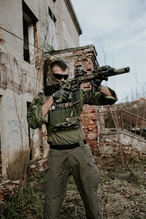 A person dressed in military-style clothing holds a rifle while standing in front of a weathered, graffiti-covered building. The individual wears sunglasses and tactical gloves, while vegetation grows around the area, indicating an abandoned or neglected setting.