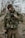 A person dressed in camouflage gear stands outdoors. The individual is equipped with tactical gear, including a vest with several attachments and protective eyewear. The background features blurred trees and foliage, suggesting a forest or woodland environment.