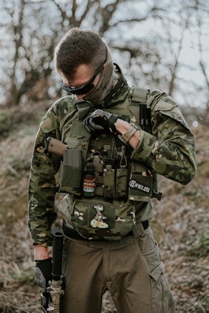 A person dressed in camouflage gear stands outdoors. The individual is equipped with tactical gear, including a vest with several attachments and protective eyewear. The background features blurred trees and foliage, suggesting a forest or woodland environment.