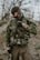 A person dressed in camouflage gear stands outdoors. The individual is equipped with tactical gear, including a vest with several attachments and protective eyewear. The background features blurred trees and foliage, suggesting a forest or woodland environment.
